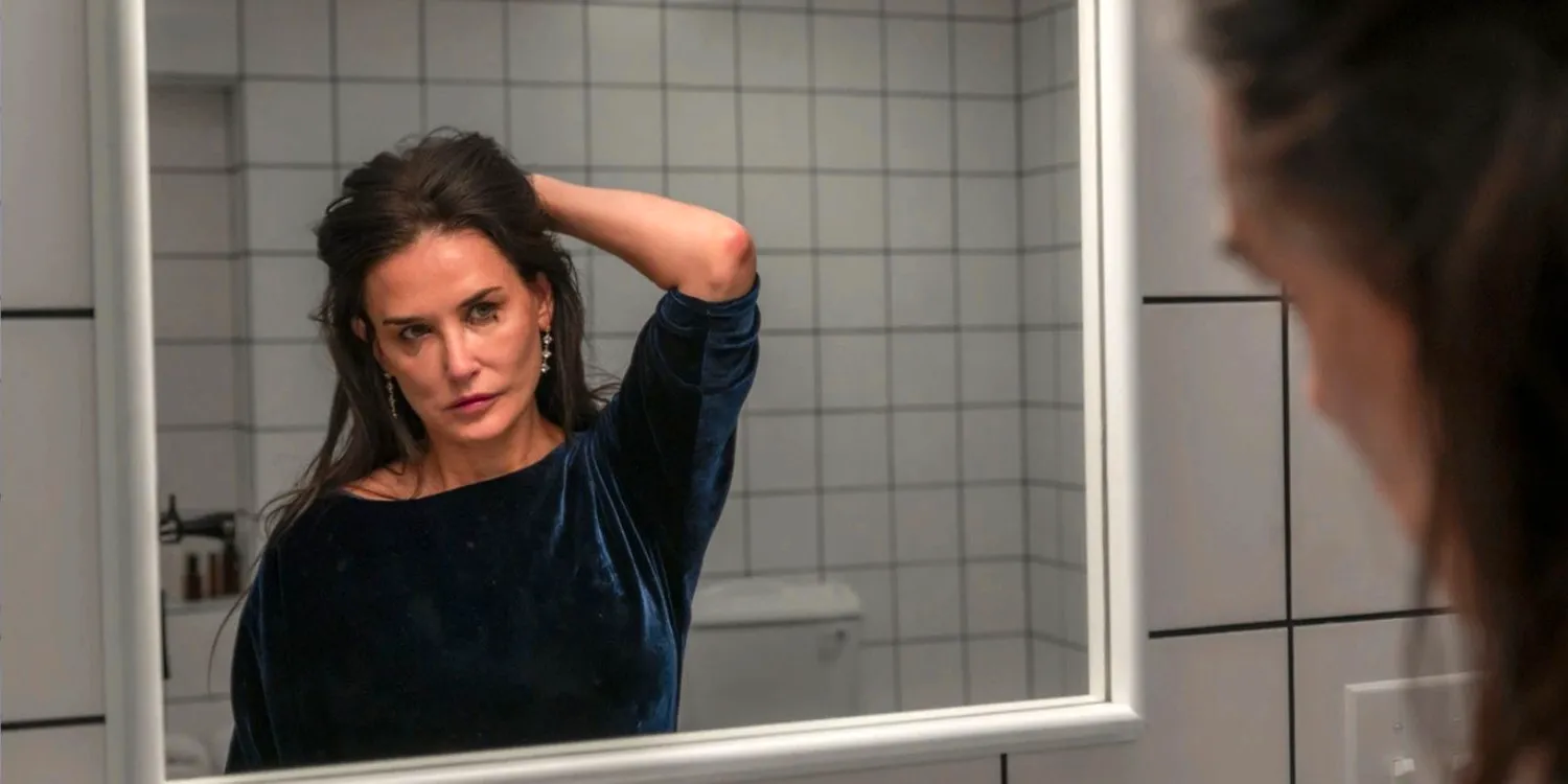 A still from The Substance. Elisabeth (played by Demi Moore) is wearing a dark blue, velvet top with a boat neckline. Holding her hair back with one hand, she regards herself in the bathroom mirror. Image