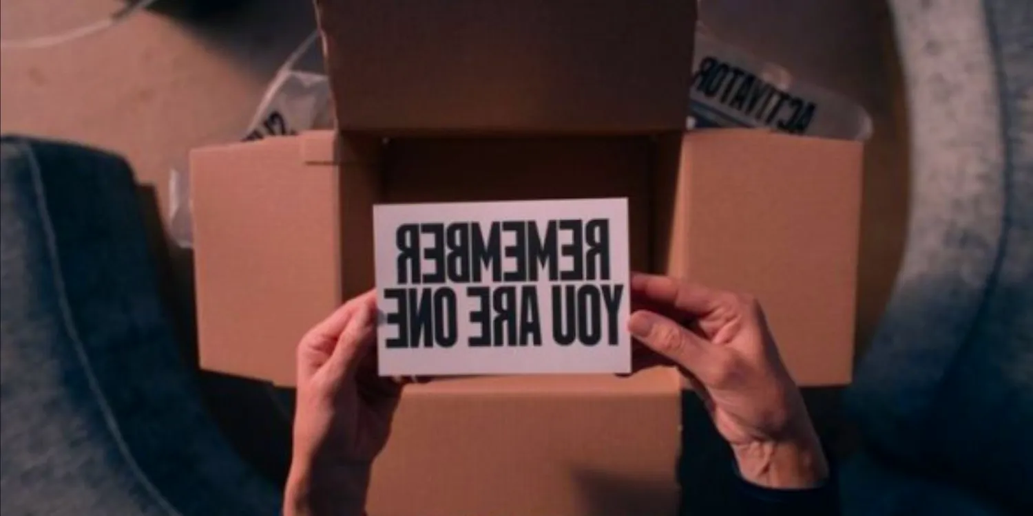 A still from The Substance. Two hands hold a note above an empty box. The note reads in graphic all-caps lettering, 'REMEMBER YOU ARE ONE'. Image