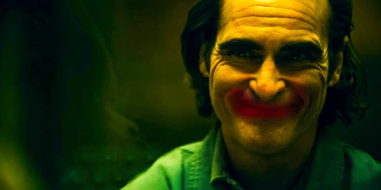 Arthur Fleck (Joaquin Phoenix) with a forced smile perfectly aligns with a red lipstick-drawn smile on a glass in Joker: Folie à Deux Image