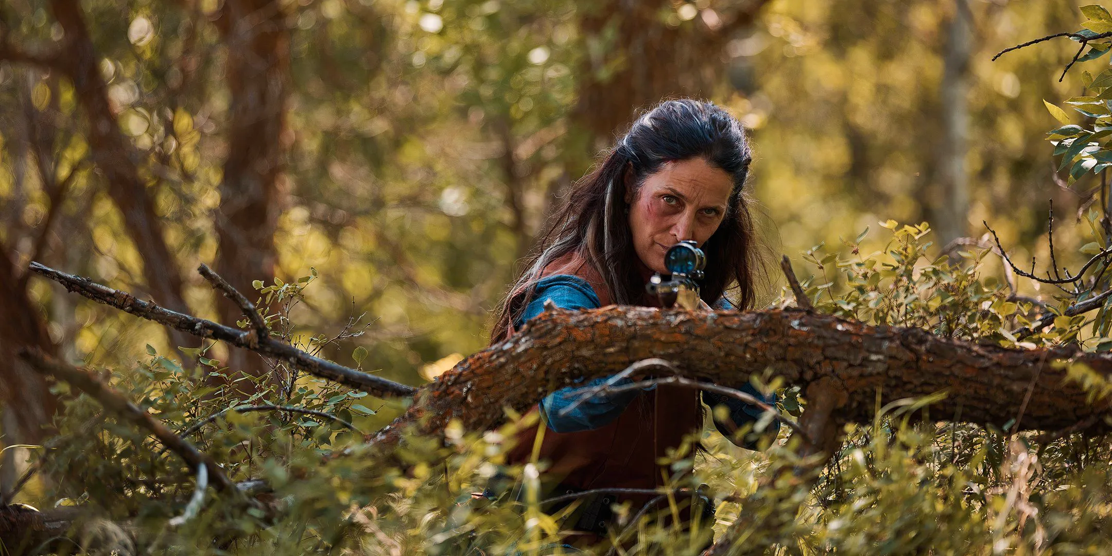 Carrie-Anne Moss as Mae aiming a sniper rifle while standing behind a tree branch in Die Alone Image