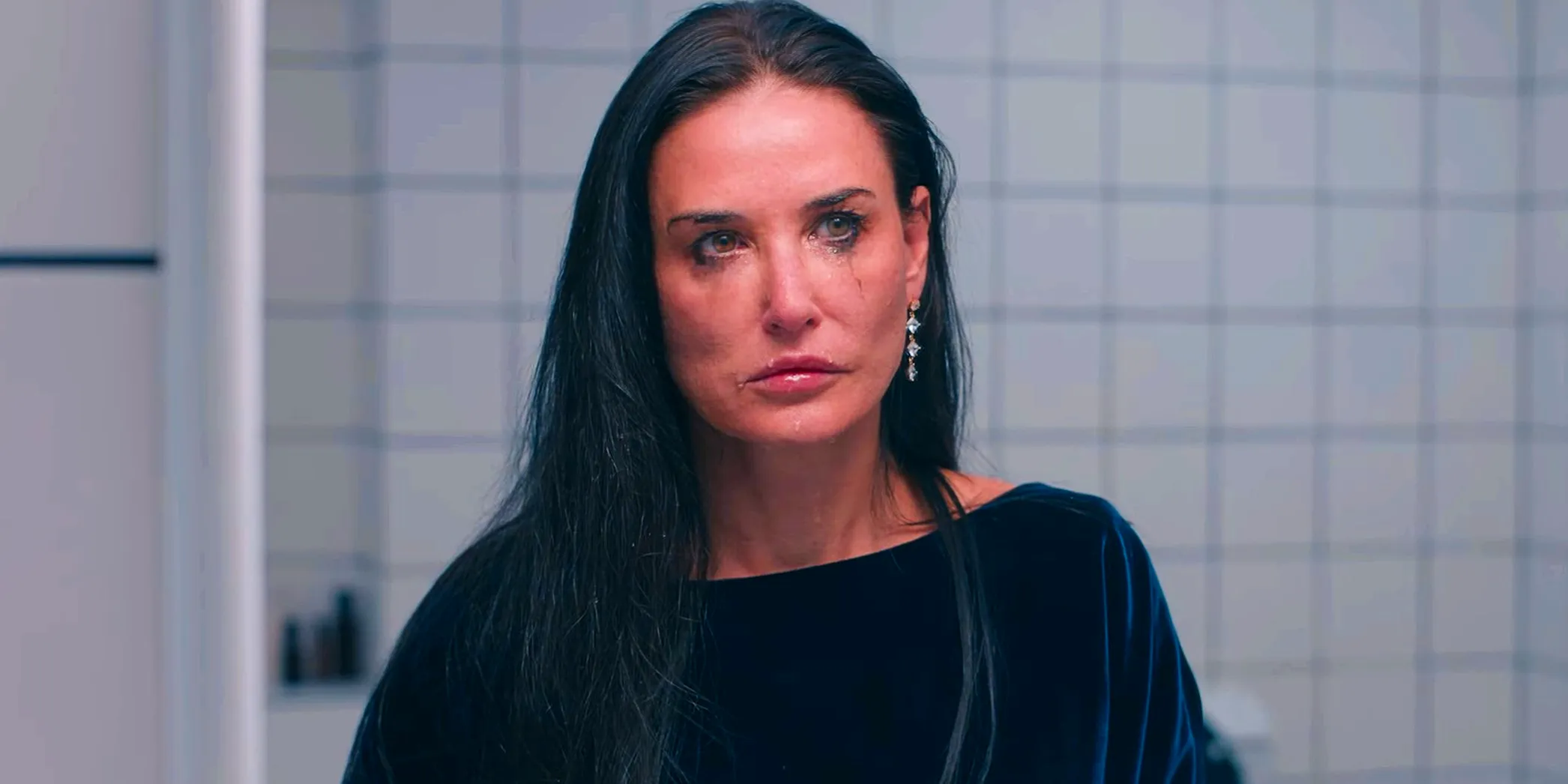 Demi Moore grimacing in the mirror of a white tiled bathroom in The Substance Image