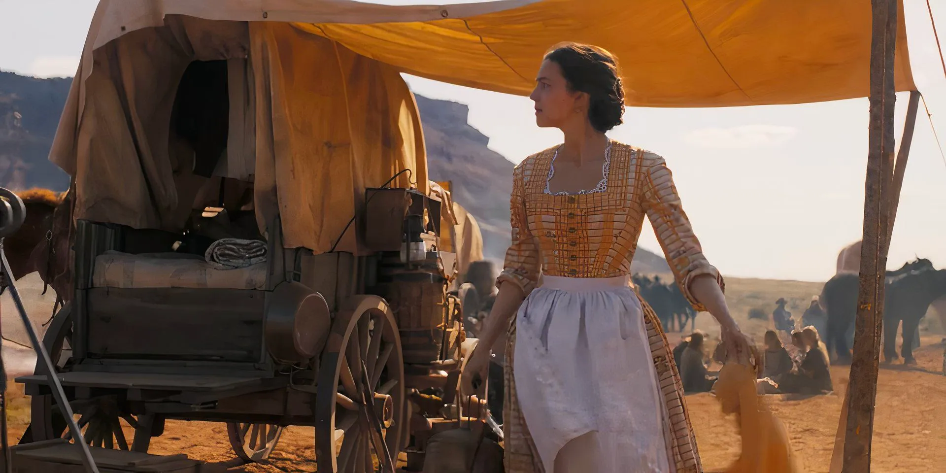 Ella Hunt as Juliette Chesney wearing an apron and holding a cloth next to a covered wagon in Horizon: An American Saga - Chapter 1 Image