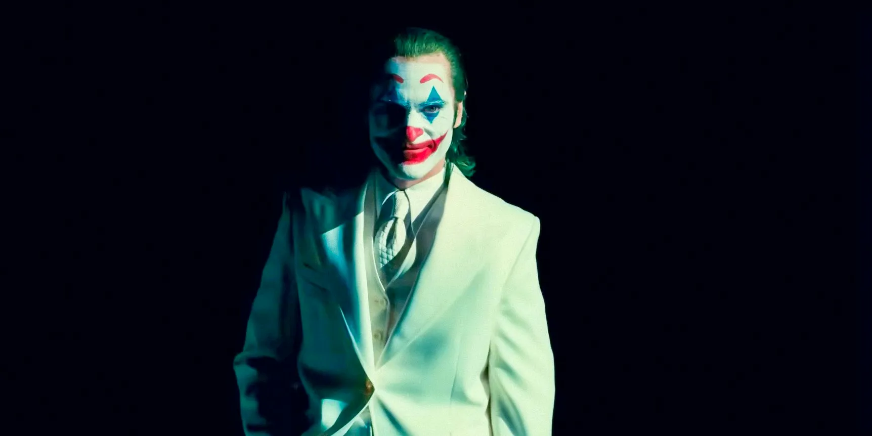 Joaquin Phoenix as Arthur Fleck/Joker wearing a white suit emerging from the darkness in Joker: Folie à Deux Image