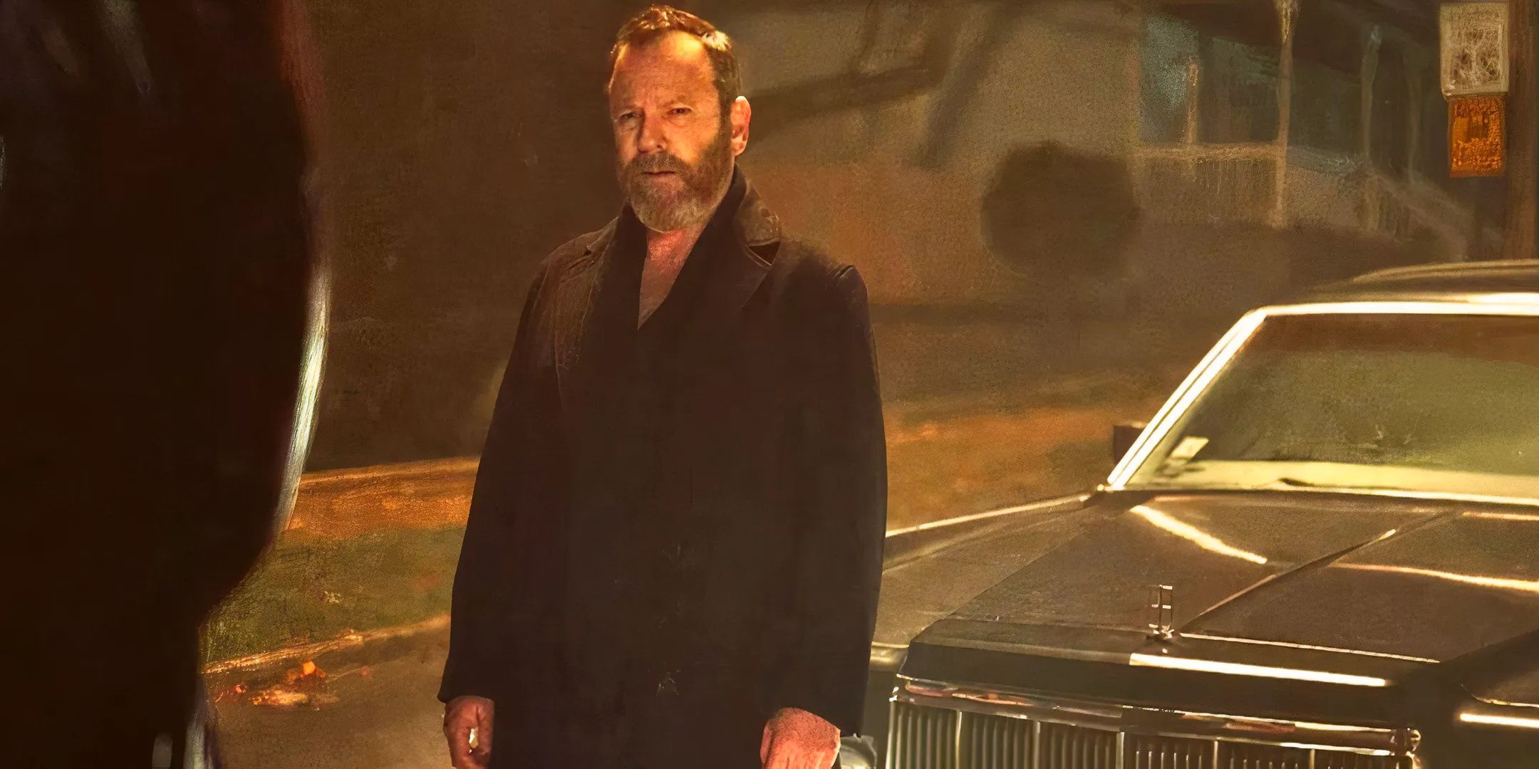 Kiefer Sutherland is standing in front of a car and squints at the people in front of him. Image