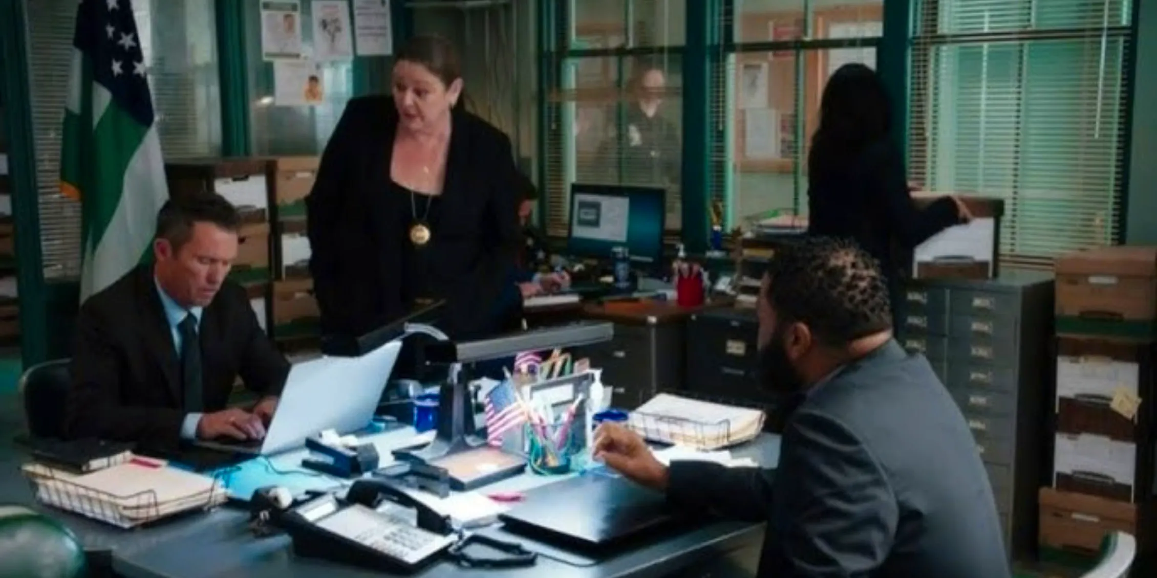 Law & Order Dixon stands at a desk giving instructions to Bernard who is sitting across from Cosgrove Image