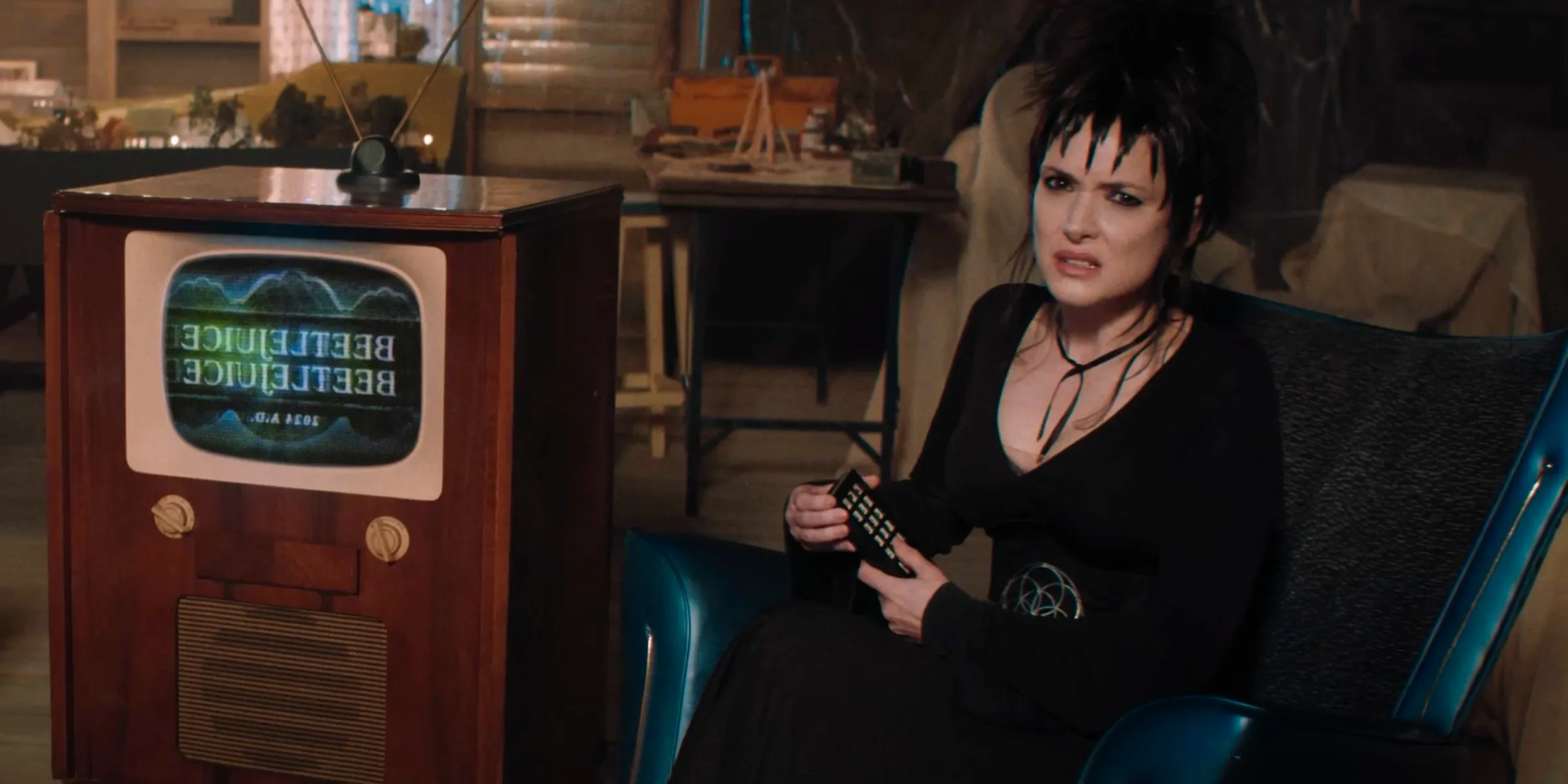 Lydia Deetz (Winona Ryder) holding a remote and sitting by a tv with the Beetlejuice 2 logo on it Image