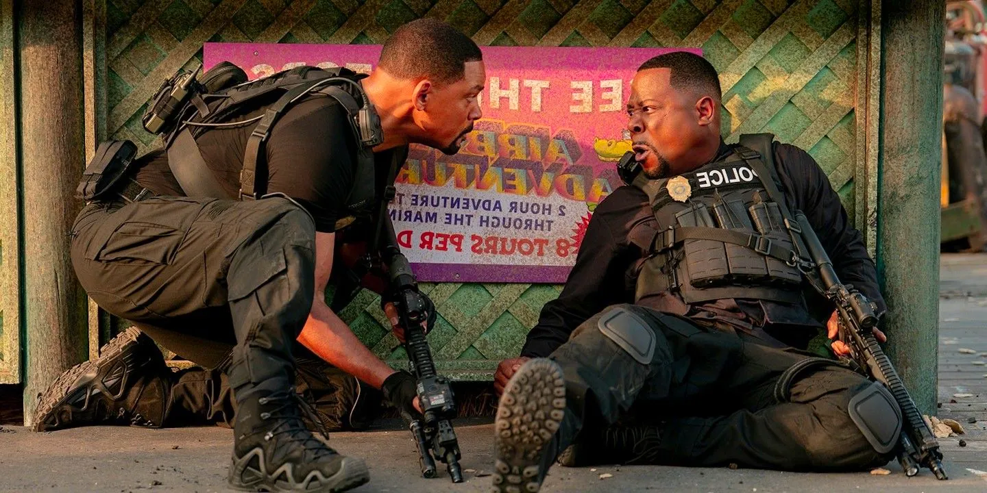 Marcus, sitting on the ground while leaning against a wall, yells at Mike, who crouches beside him holding a gun in Bad Boys Ride or Die Image