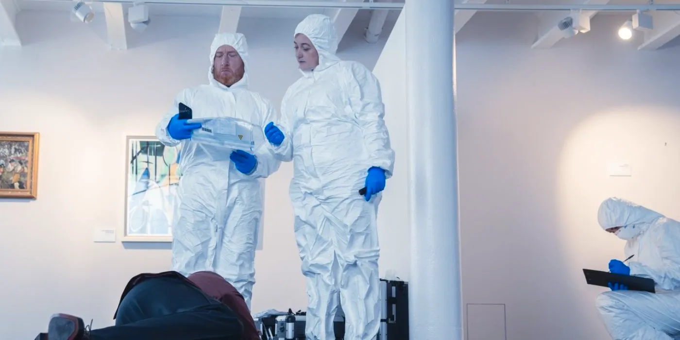 Max (Adrian Scarborough) standing over a body in an art gallery dressed in a protective suit with another woman in The Chelsea Detective. Image