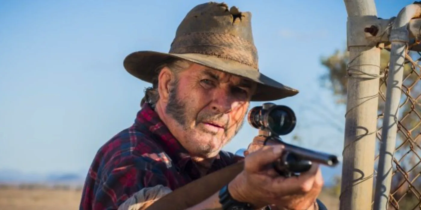 Mick Taylor aims his gun at someone while standing next to a fence in Wolf Creek 2 (2013) Image