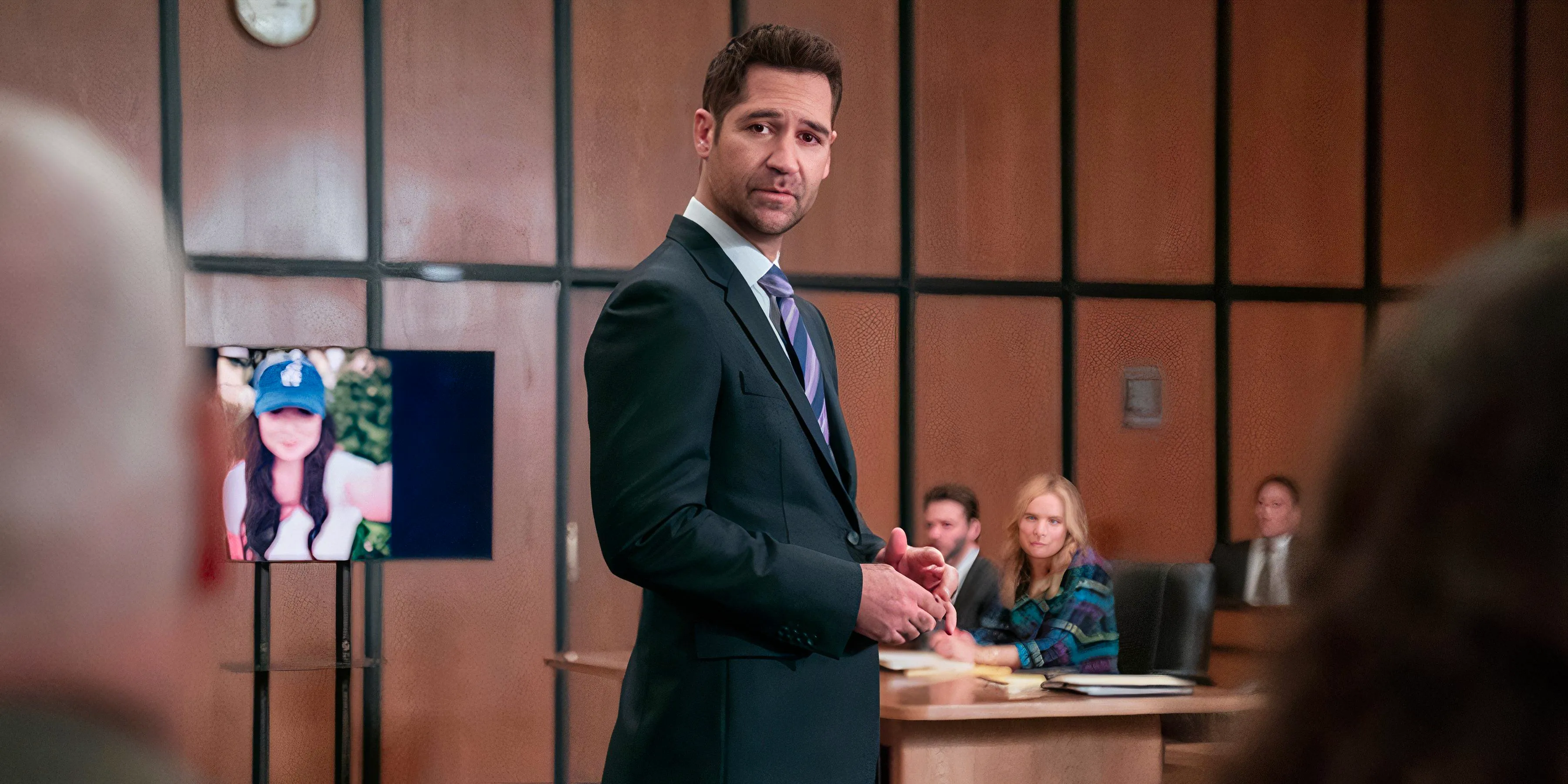 Mickey stands in the courtroom with a picture of Glory Days in the background in The Lincoln Lawyer season 3 episode 6 Image