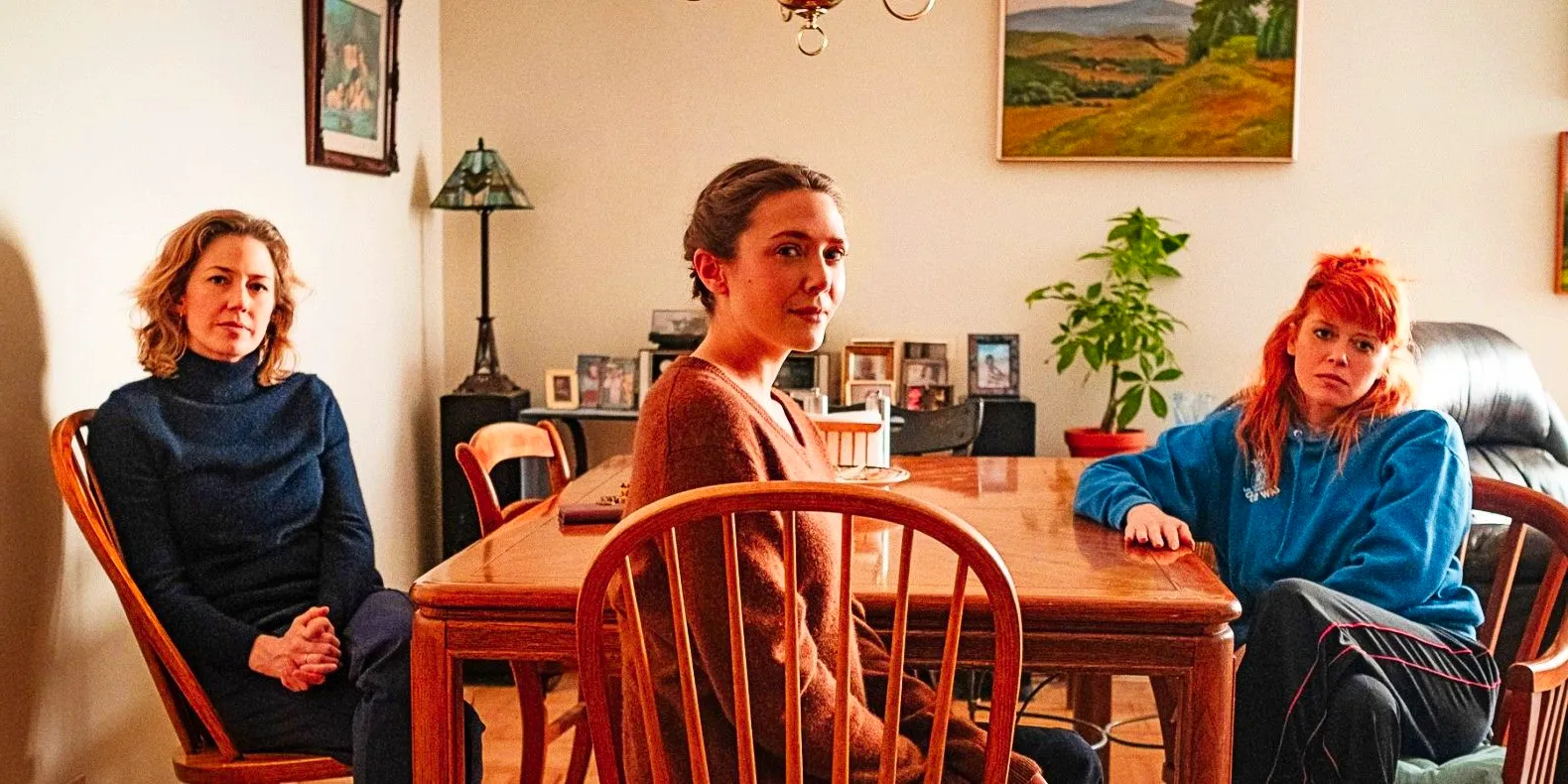 Natasha Lyonna, Elizabeth Olsen, and Carrie Coon sit at a kitchen table and look toward the camera in His Three Daughters Image