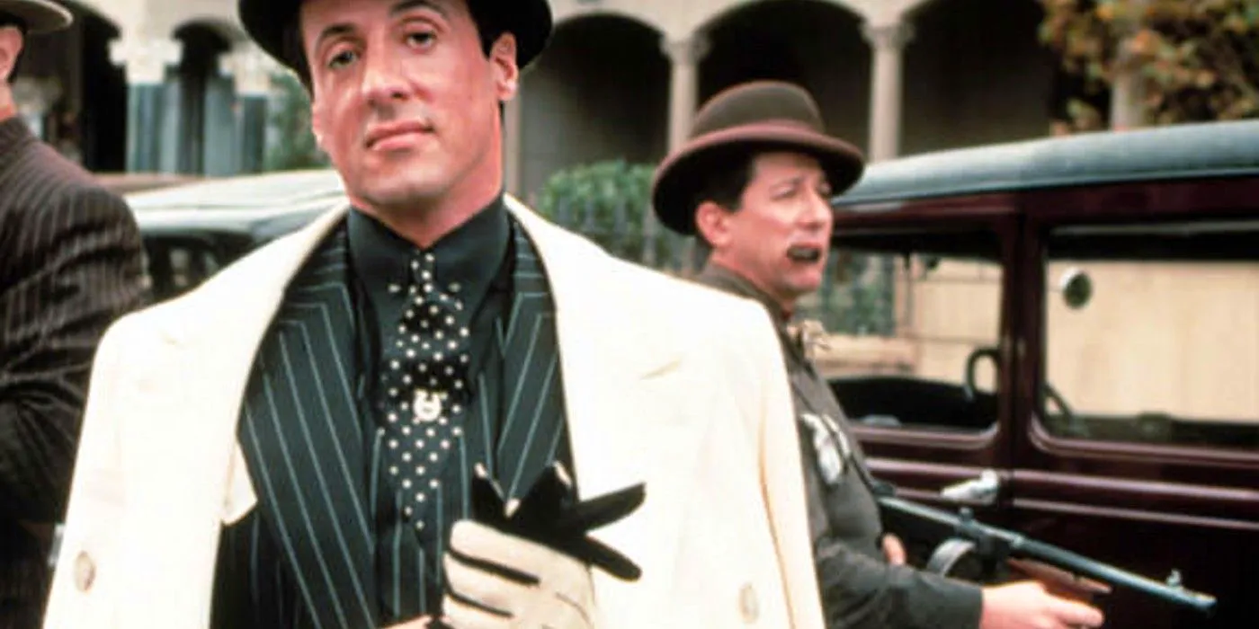 Sylvester Stallone in Oscar, wearing a white coat with a dark suit and holding gloves with a henchman with a gun in the background Image