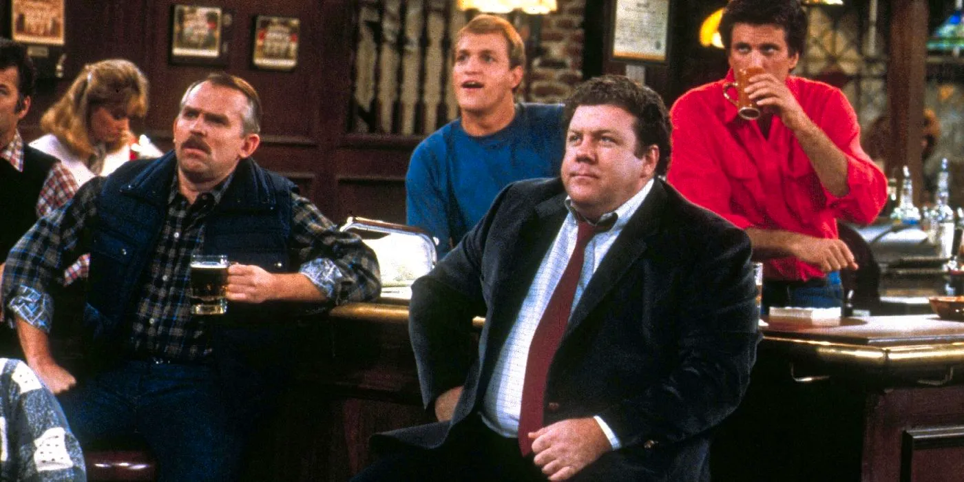 The cast of Cheers sits at the bar looking at something off-screen. Image