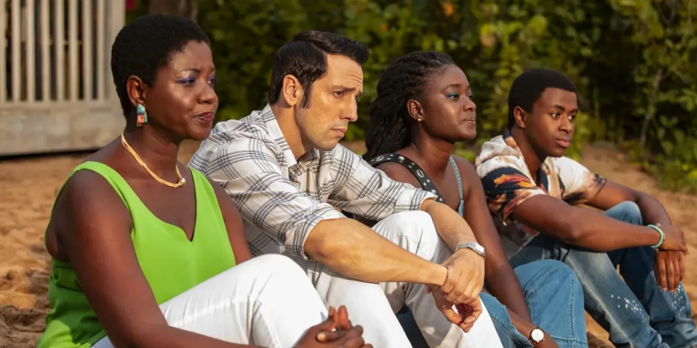 The cast of Death in Paradise sitting on the beach and looking upset. Image