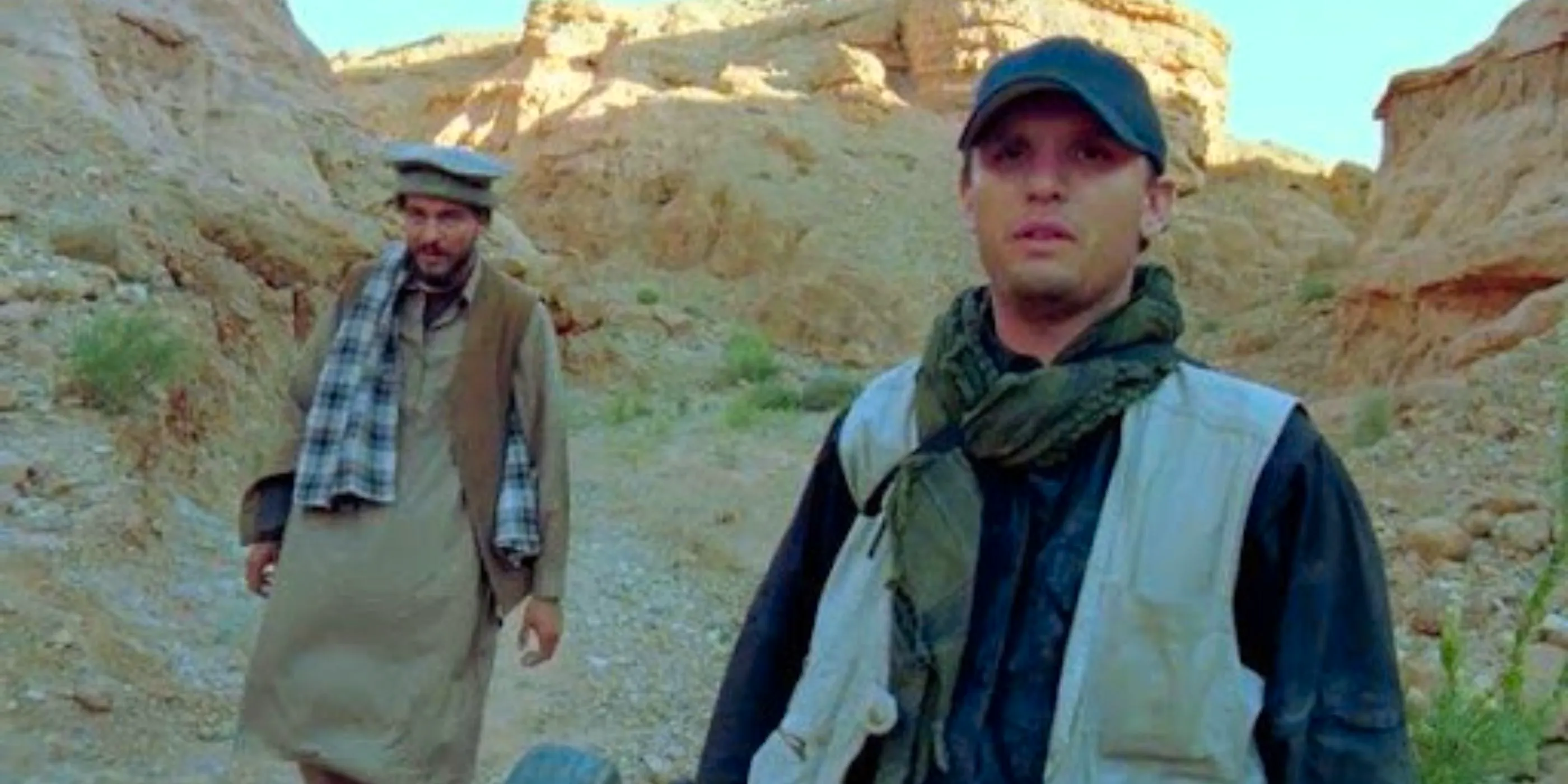 Two men in desert clothing are stood in a desert by some hills staring ahead intently in The Objective Image