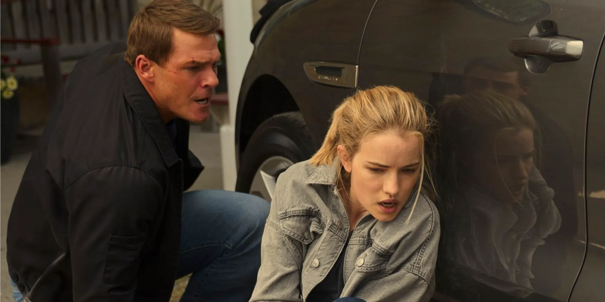 Willa Fitzgerald as Roscoe Conklin hiding behind a car with Alan Ritchson's Jack Reacher. Image