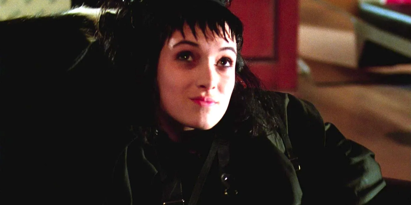 Winona Ryder as Lydia Deetz in Beetlejuice sitting in a chair all dressed in black with a pale face and jagged bangs, looking up sort of smugly. Image